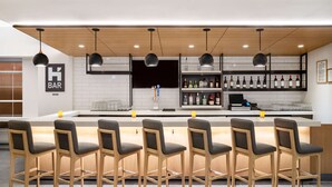 Zona de bar moderna con taburetes de madera, un revestimiento de baldosas blancas y una variedad de botellas de licores en estanterías.