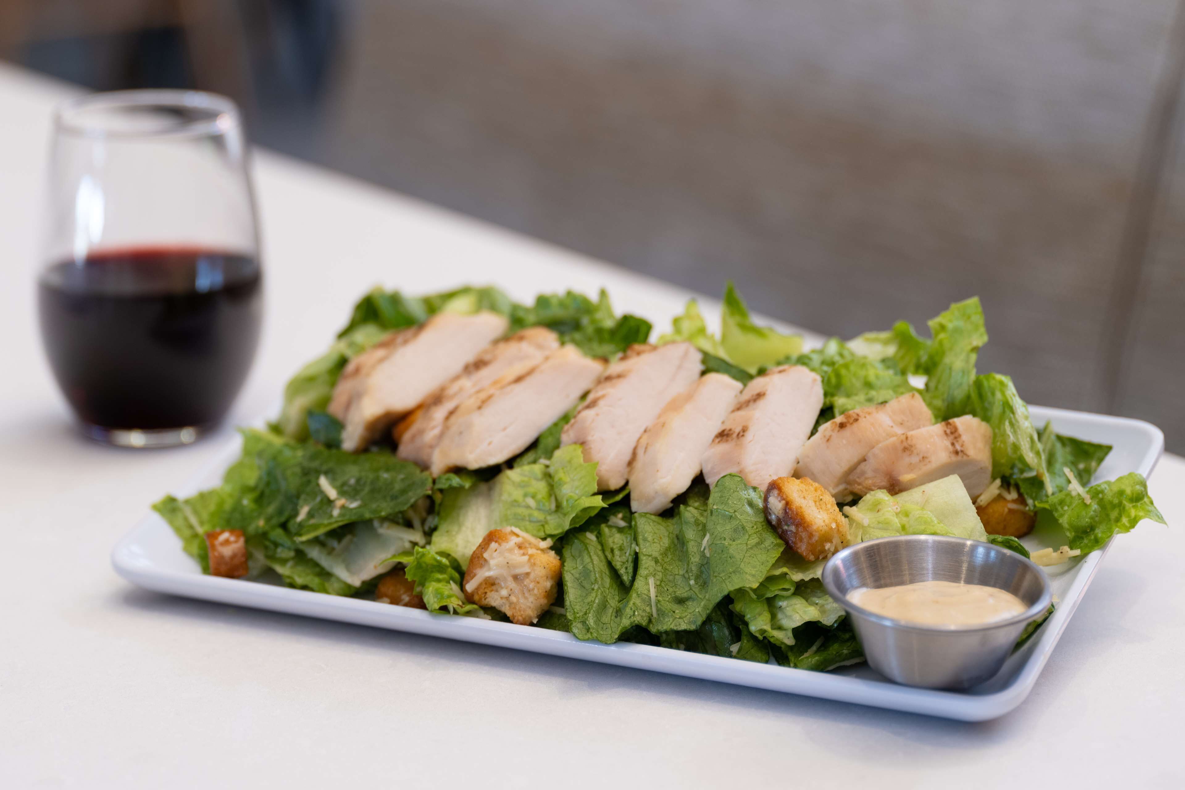 A plate of chicken salad with croutons and a small bowl of dressing, accompanied by a glass of red wine.