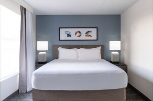 Una habitación de hotel con una cama grande, dos lámparas de noche, una obra de arte enmarcada sobre la cama y una ventana con cortinas.
