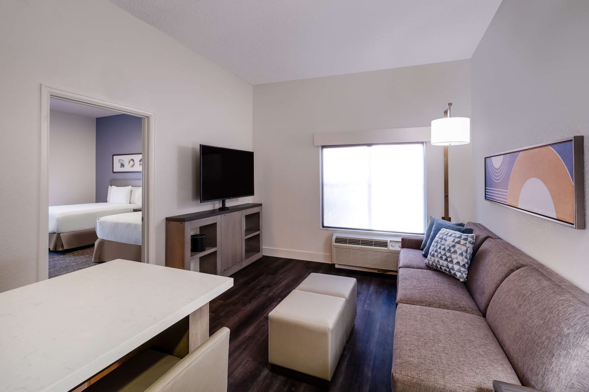 A modern hotel room with a sofa, a TV, a dining table, and a window with a view.