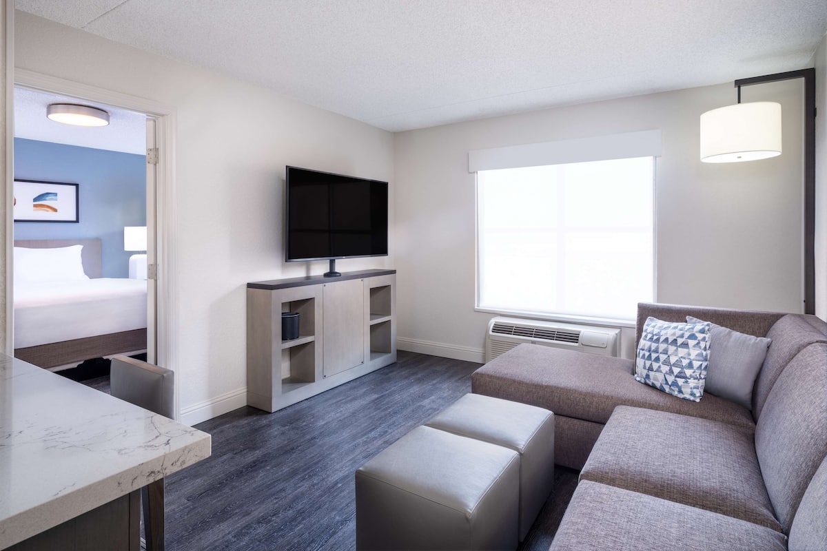 A modern hotel room with a flat-screen TV, a sofa with a geometric pillow, a dining area with a marble countertop, and a large window providing natural light.