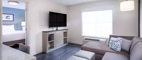 A modern hotel room with a flat-screen TV, a sofa with a geometric pillow, a dining area with a marble countertop, and a large window providing natural light.