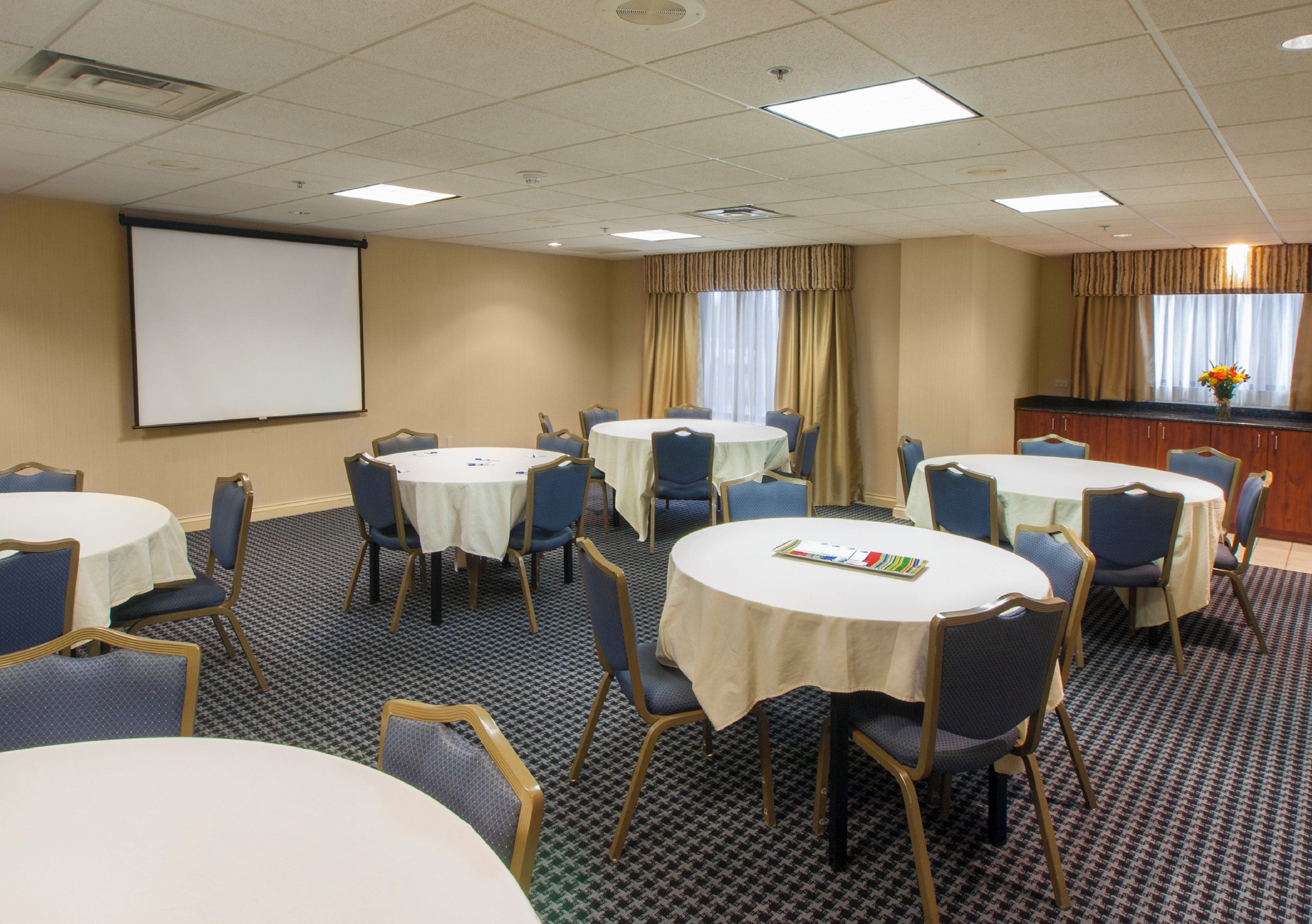 A conference room with multiple tables and chairs, a projector screen, and a window with curtains.