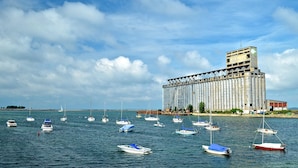 A large industrial building with a silo-like structure and a tower on top, surrounded by calm water with several small boats.