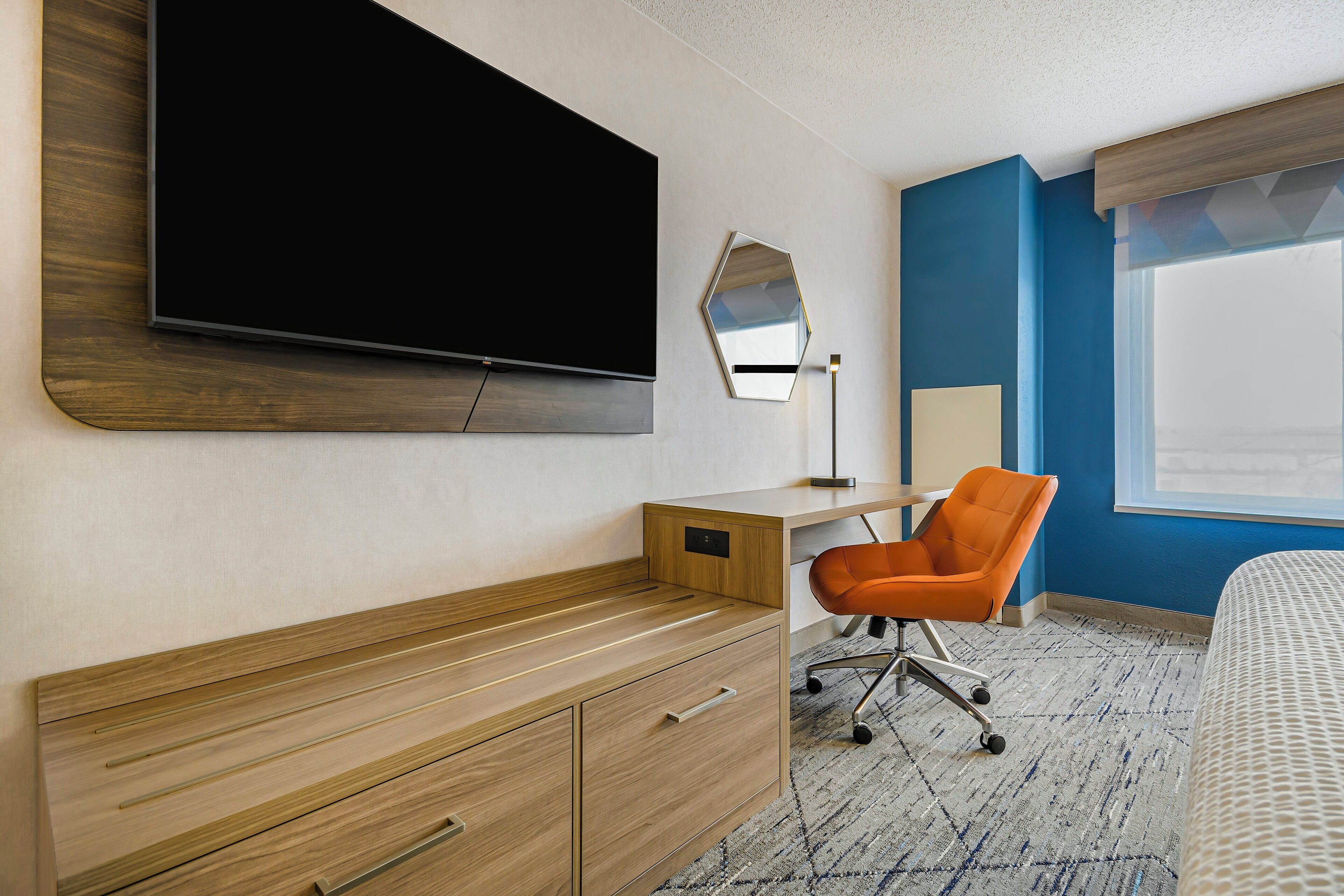 Habitación de hotel con escritorio de madera, una silla naranja, un televisor de pantalla plana en la pared, un espejo y una cama con colcha estampada.