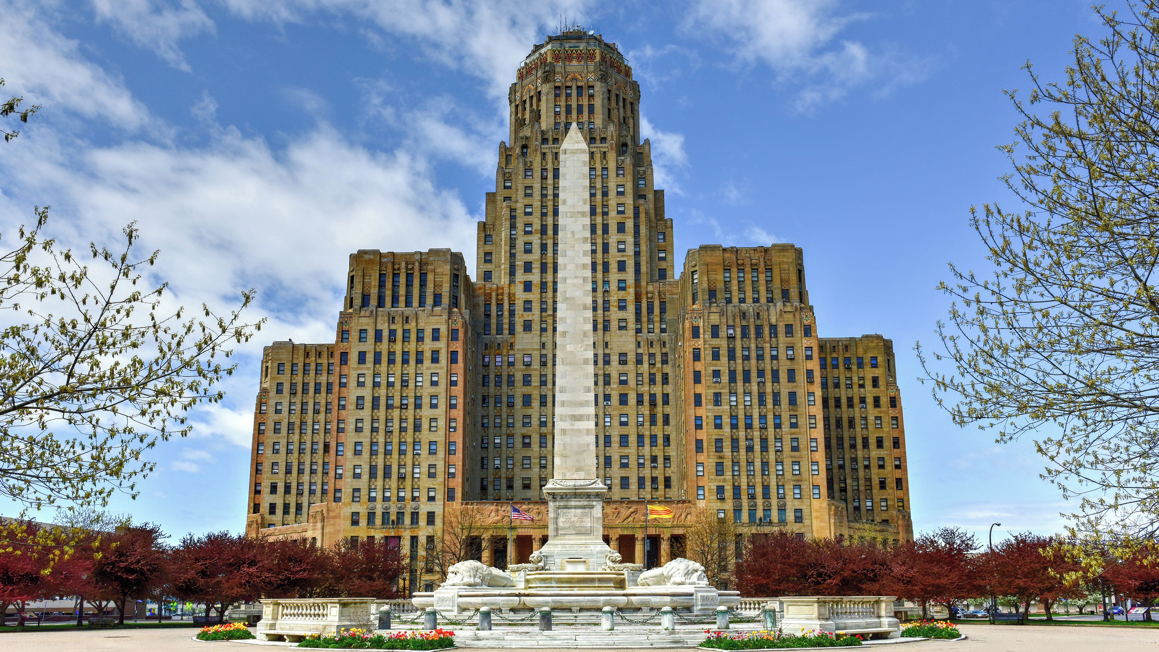 A large, historic building with a central tower and multiple smaller towers, surrounded by a landscaped area with a fountain and trees.