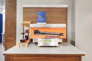 A kitchen area with a countertop displaying a pancake-making machine, a sign indicating the machine is out of action, and two bottles of syrup on a wooden stand. - Holiday Inn Express Hotel & Suites Buffalo-Airport by IHG (Buffalo)