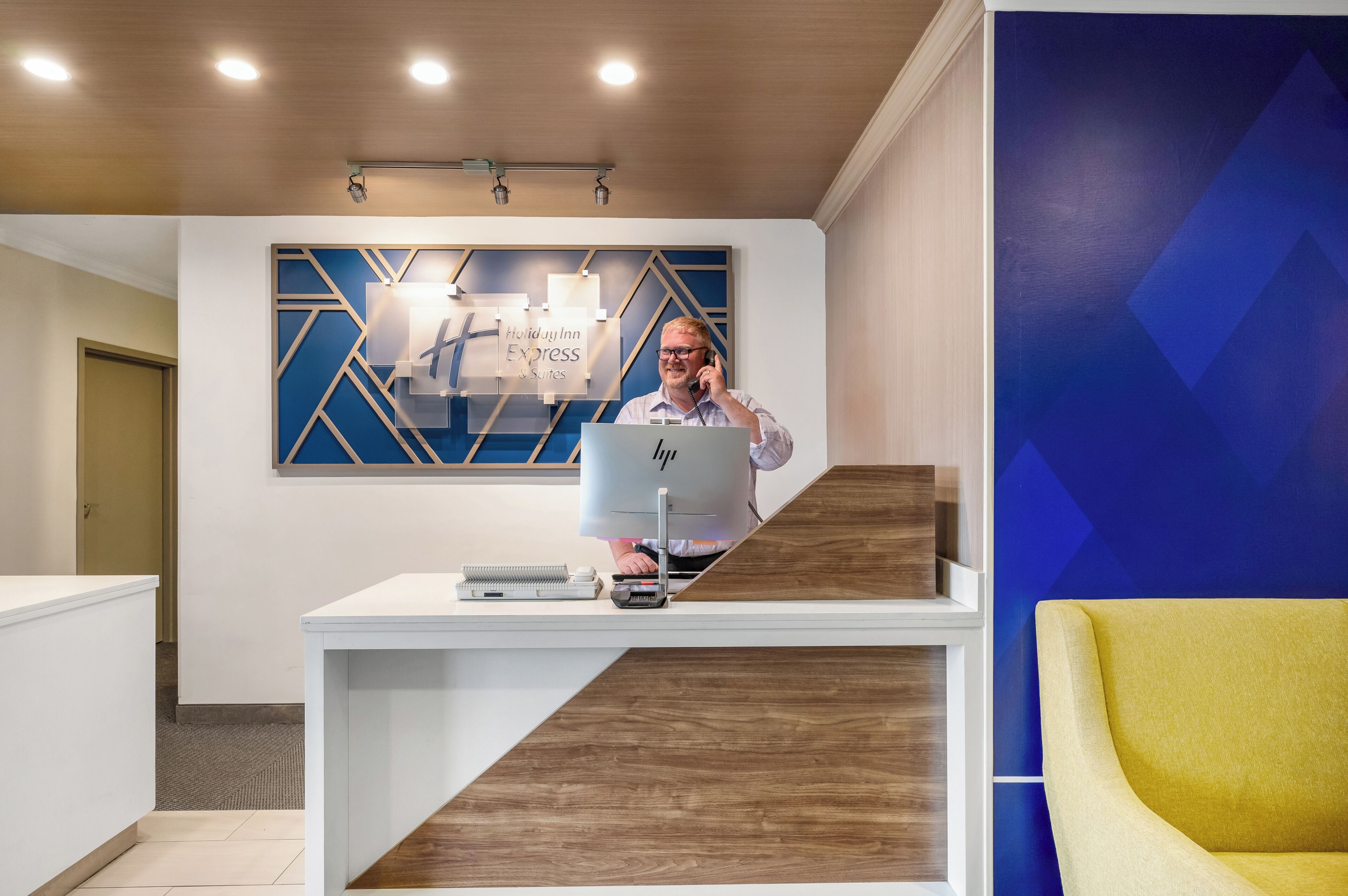 Un mostrador de recepción con una computadora, un teléfono y una caja registradora. Una pared azul con un diseño geométrico y un cartel que dice 'Holiday Inn Express & Suites'.
