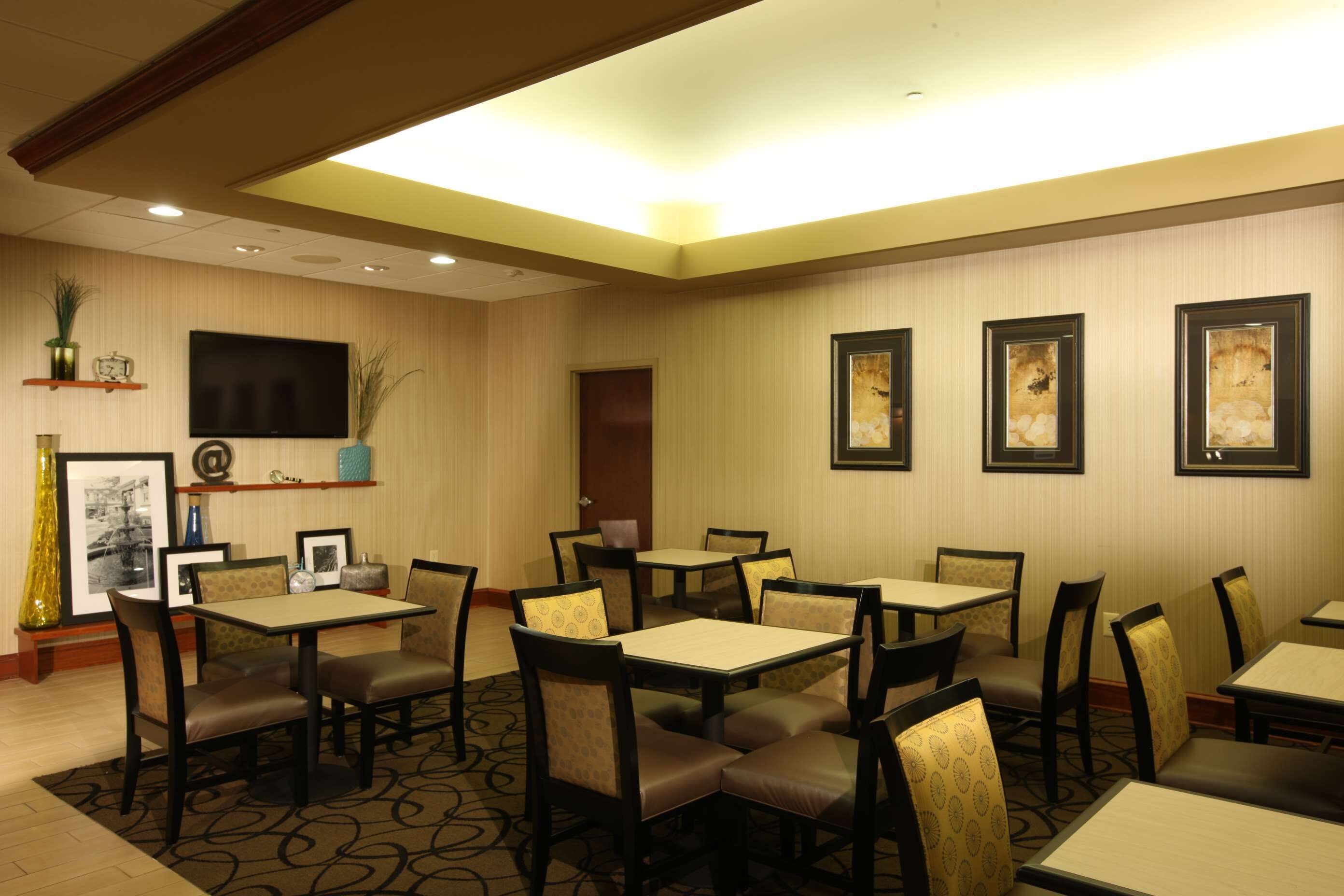 A hotel dining area with multiple tables and chairs, a flat-screen TV mounted on the wall, and framed artwork on the walls.