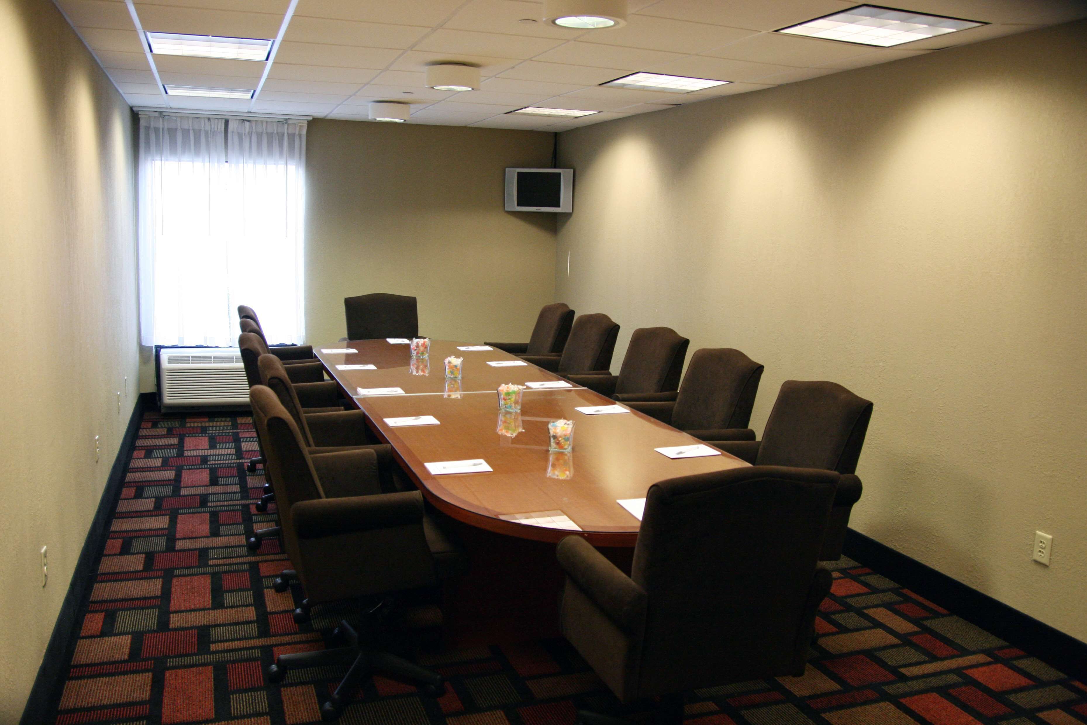 A conference room with a long table, multiple chairs, a flat-screen TV on the wall, and a patterned carpet.