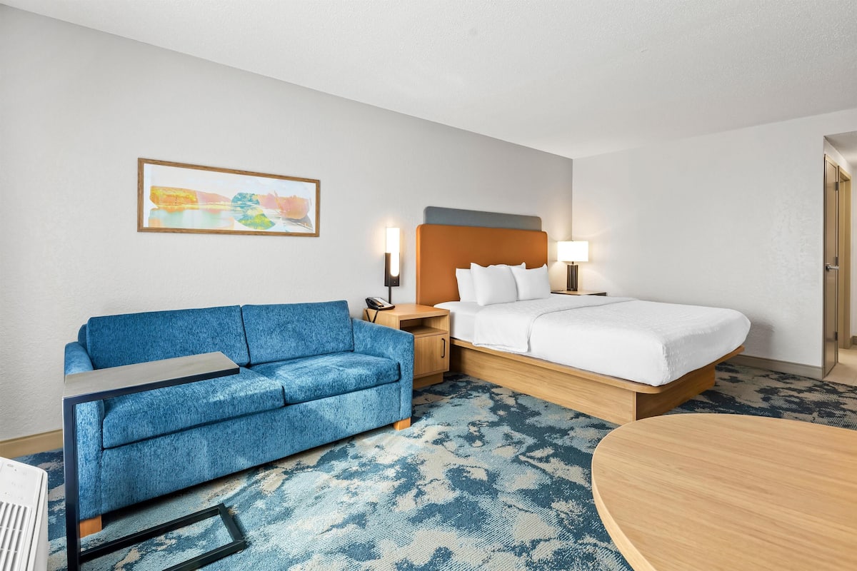 A hotel room with a blue sofa, a bed with white bedding, a wooden nightstand, a lamp, and a framed picture on the wall.