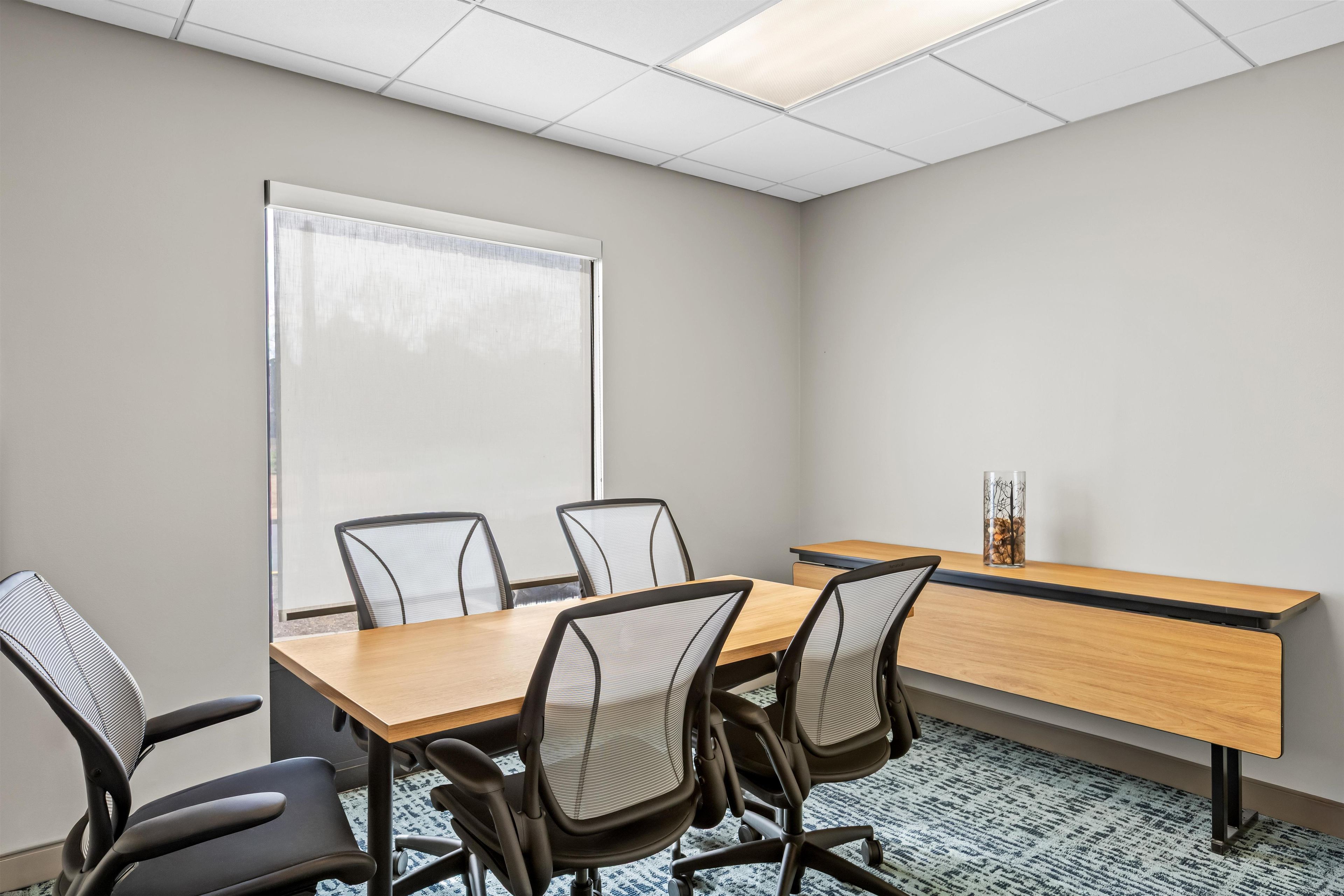 A conference room with a long table, four chairs, a whiteboard, and a window with blinds.