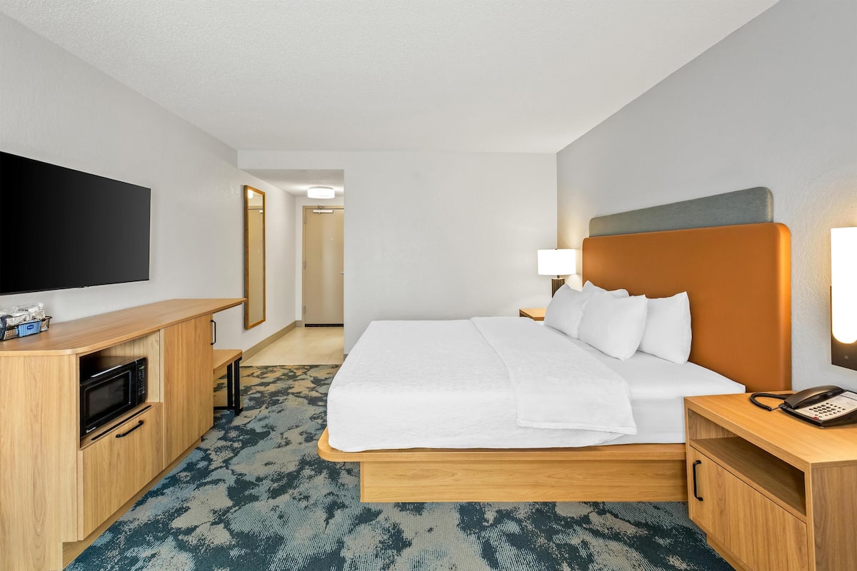 A hotel room with a bed, a TV, a desk with a microwave, and a bedside table with a lamp and phone.