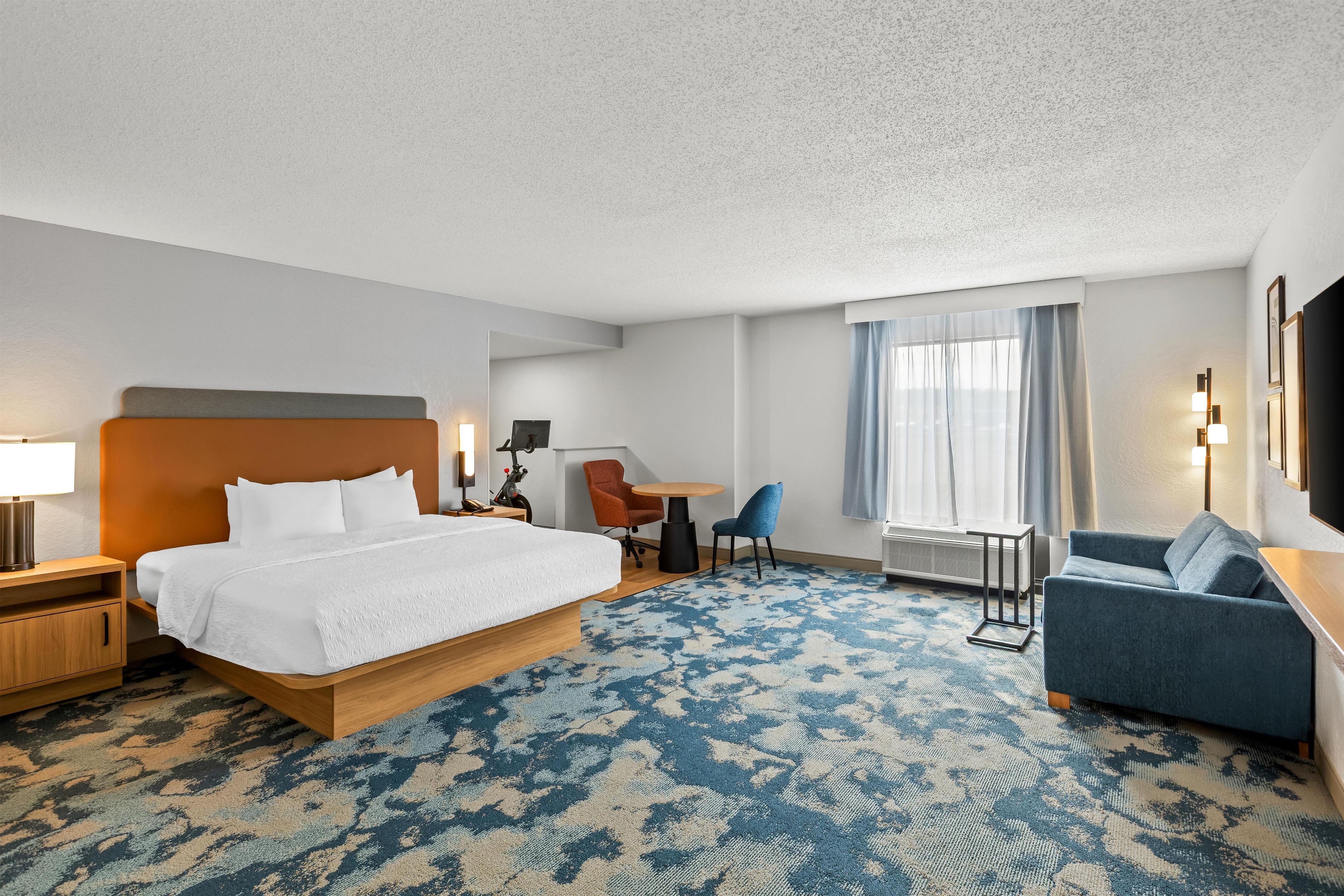 A hotel room with a bed, a desk, a chair, a sofa, and a large window with curtains.