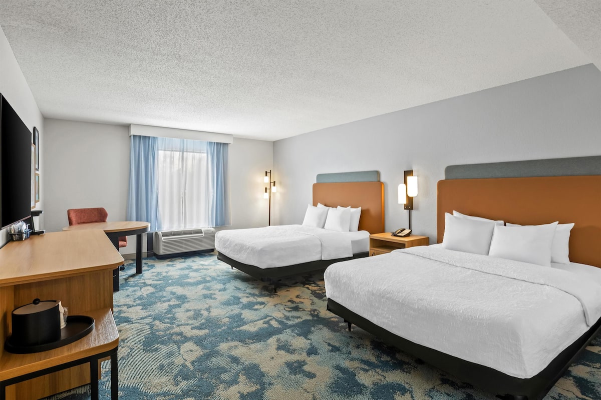 A hotel room with two beds, a desk, a chair, a TV, and a window with curtains.