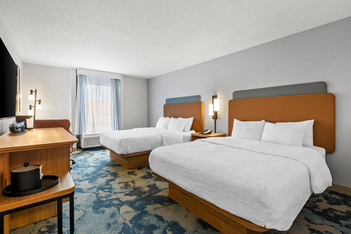 A hotel room with two beds, a desk, a chair, a flat-screen TV, and a window with curtains.
