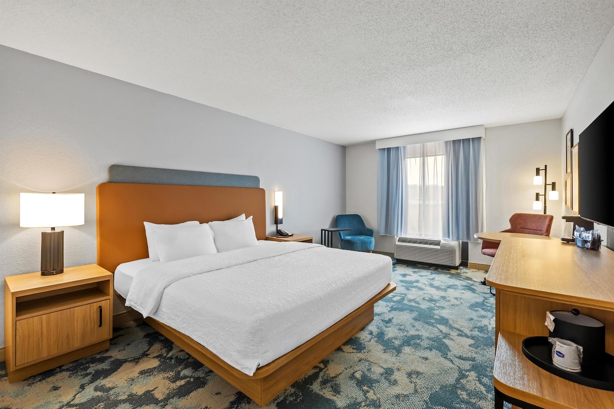 A hotel room with a large bed, a desk, a chair, a TV, and a window with curtains.