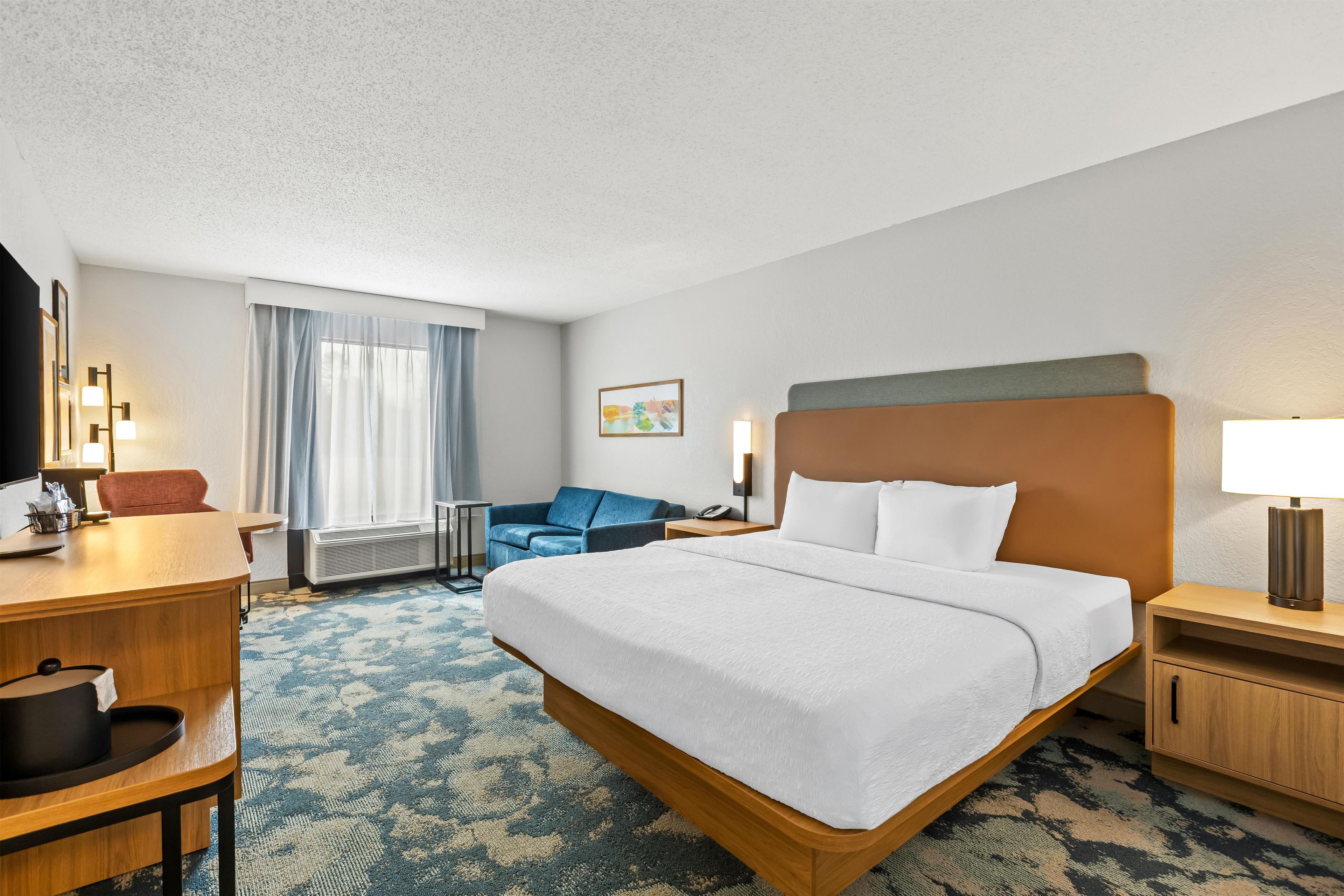 A hotel room with a bed, a desk, a chair, a desk lamp, a nightstand, a wall-mounted lamp, a window with curtains, and a framed picture on the wall.