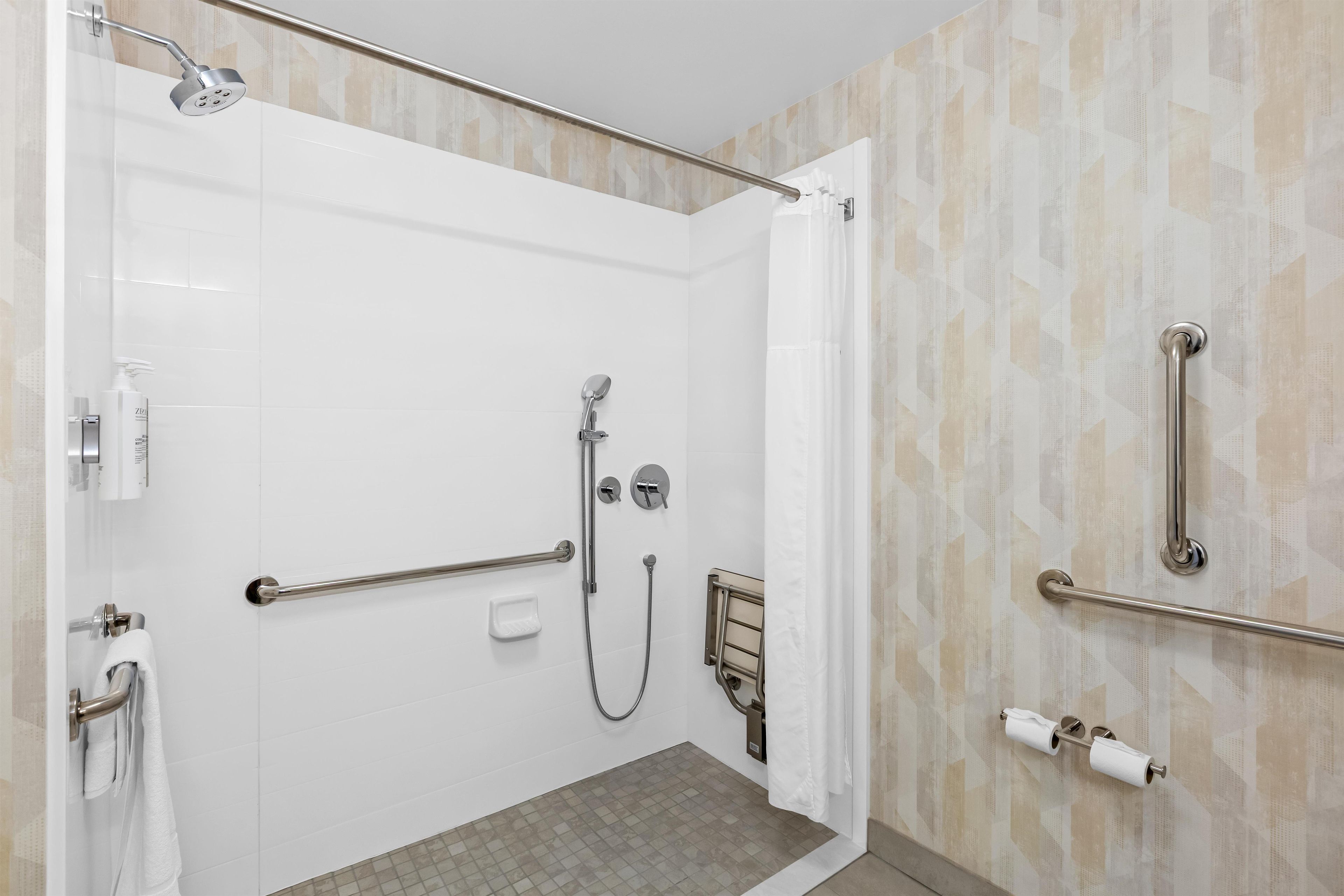A modern shower area with a glass door, grab bars, a handheld showerhead, and a handheld shower attachment.