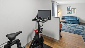 A room with a stationary bike, a blue sofa, a small table, and a TV mounted on the wall. - Hampton Inn Carrollton (Carrollton)