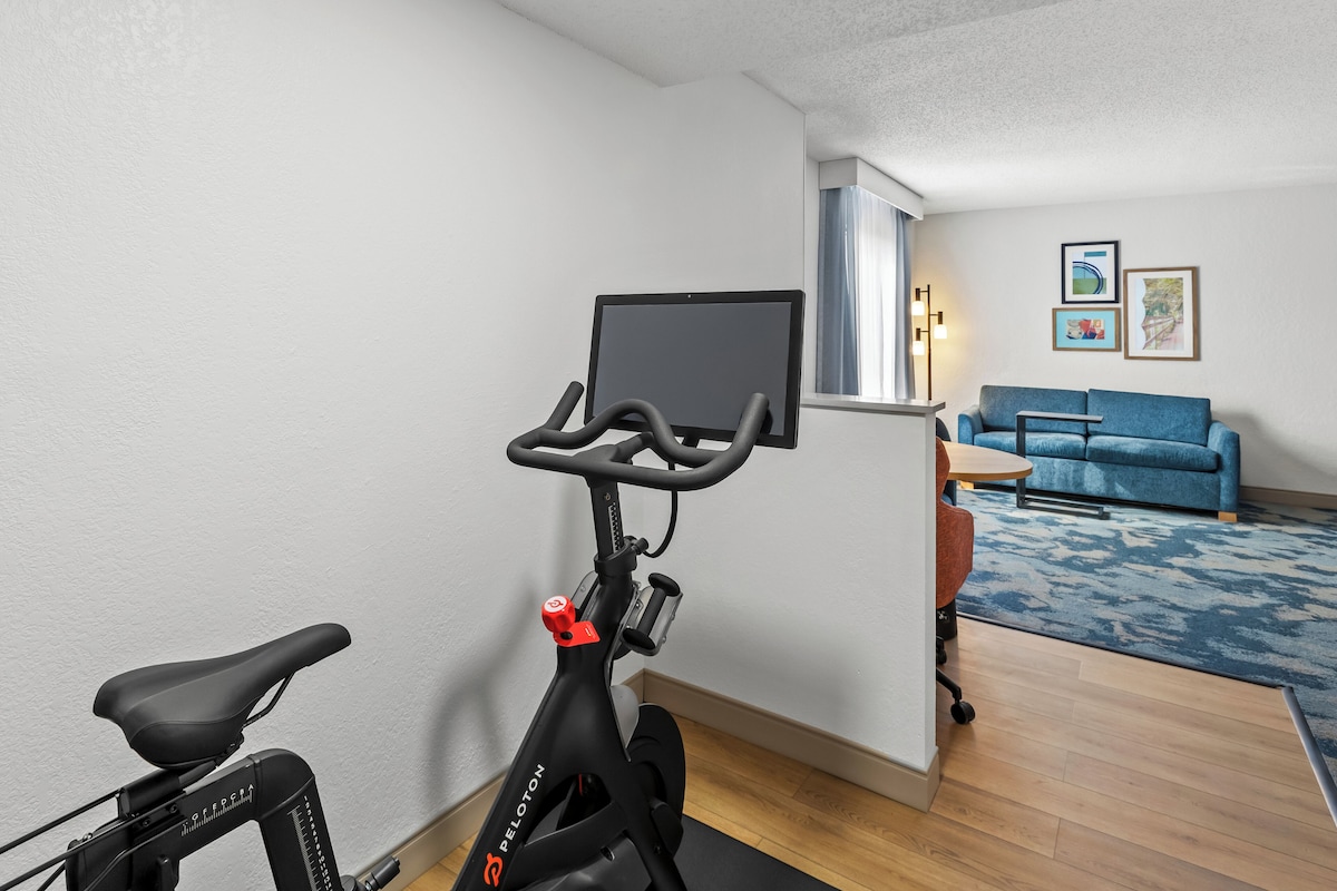Una stanza con una cyclette, un divano blu, un tavolino e una TV montata a parete.