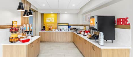 A clean, modern breakfast area with a central counter, various food and drink dispensers, and a sign for The Hampton Waffle.