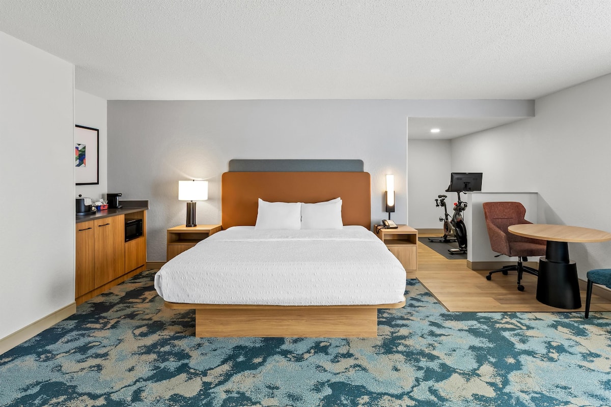 A hotel room with a large bed, wooden headboard, bedside tables with lamps, a desk with a chair, and a small kitchenette.