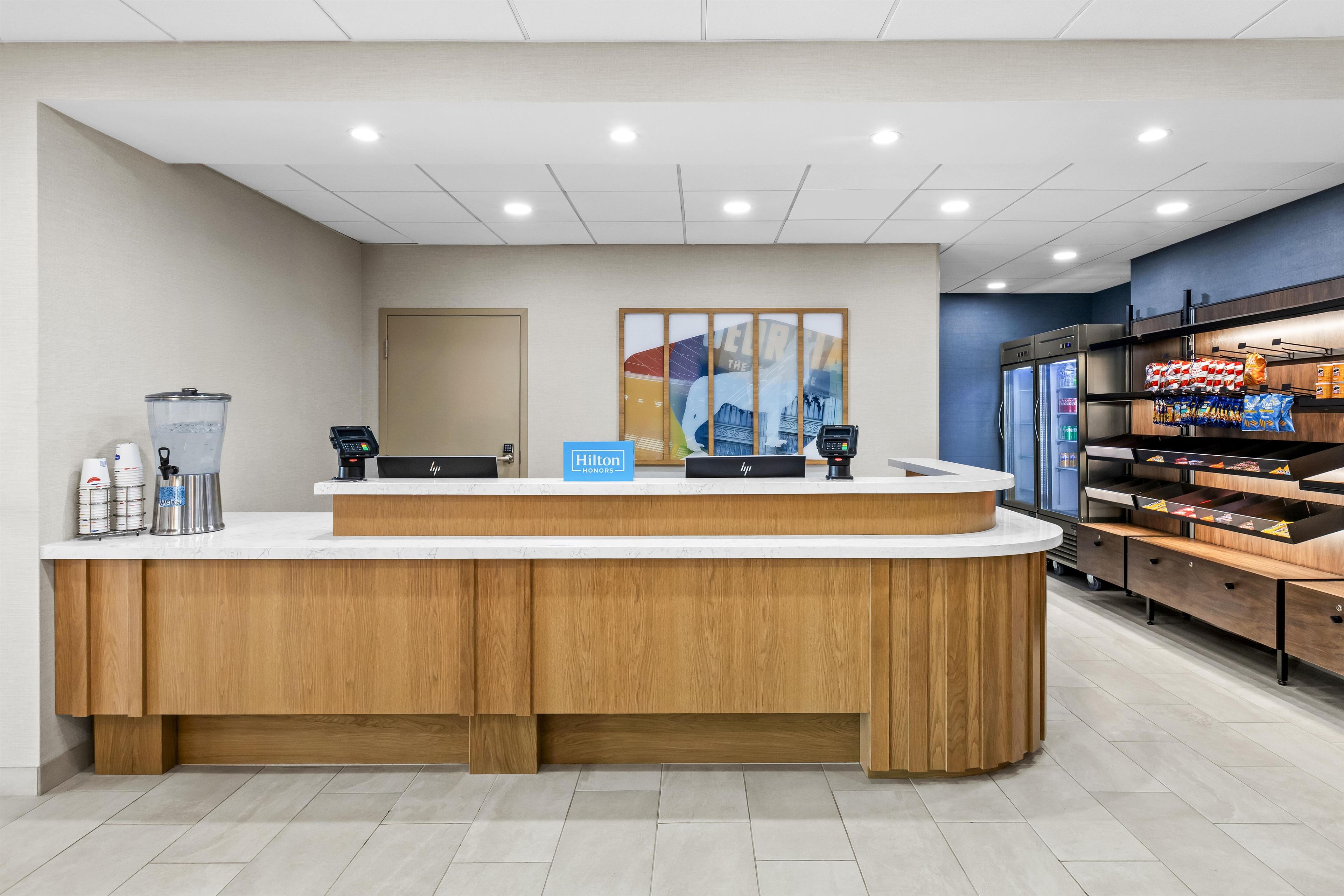 A hotel reception area with a wooden reception desk, a water dispenser, a coffee machine, and a minibar.