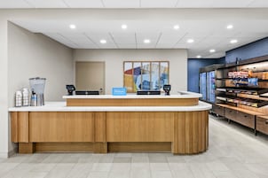 Zona de recepción de hotel con mostrador de madera, dispensador de agua, máquina de café y minibar.
