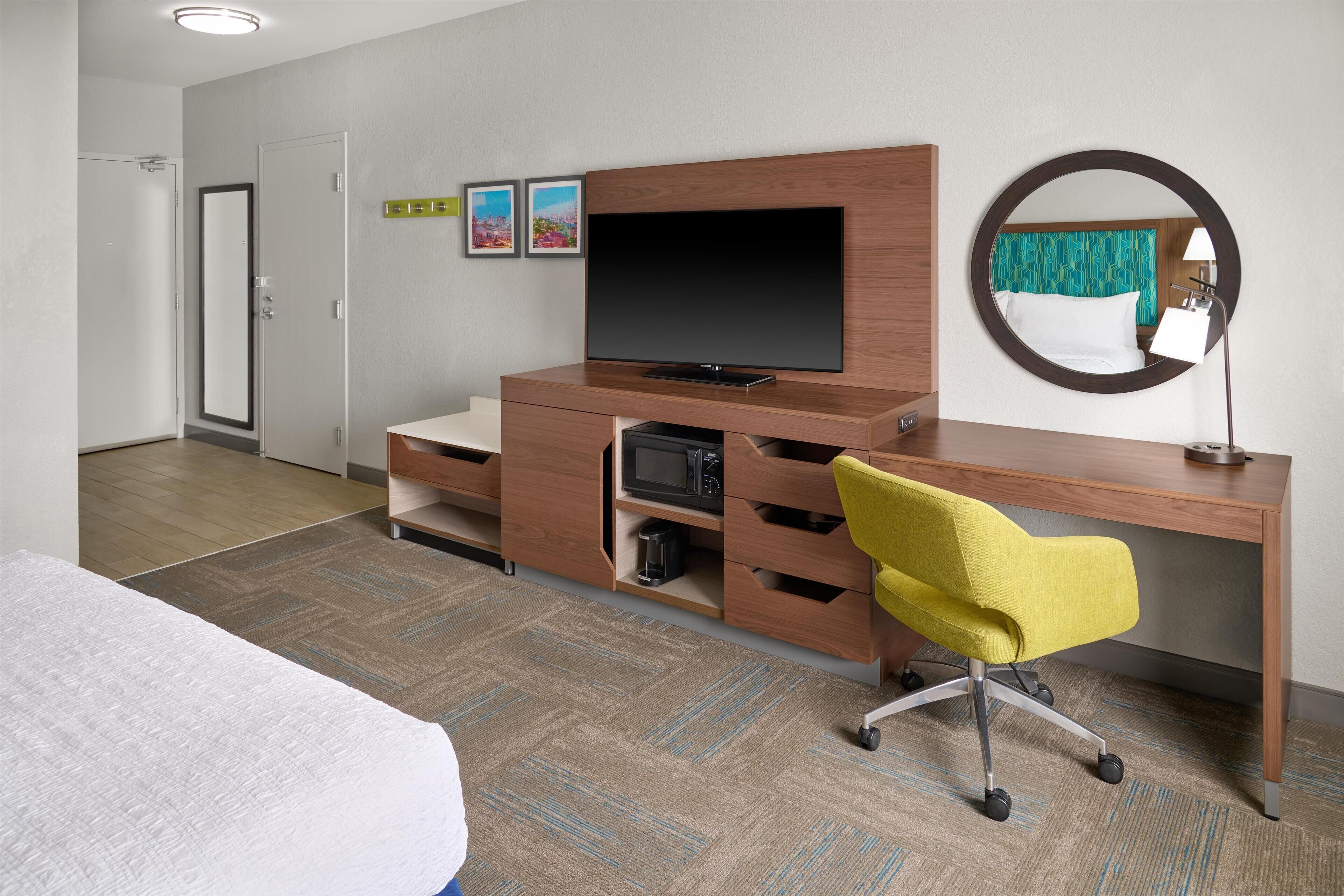 Una habitación de hotel con una cama, un escritorio de madera con un televisor, una silla amarilla, un microondas y un espejo en la pared.