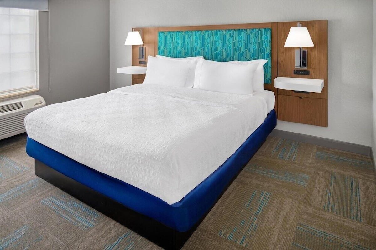 A hotel room with a large bed, two bedside tables with lamps, a headboard with a patterned design, and a window with blinds.