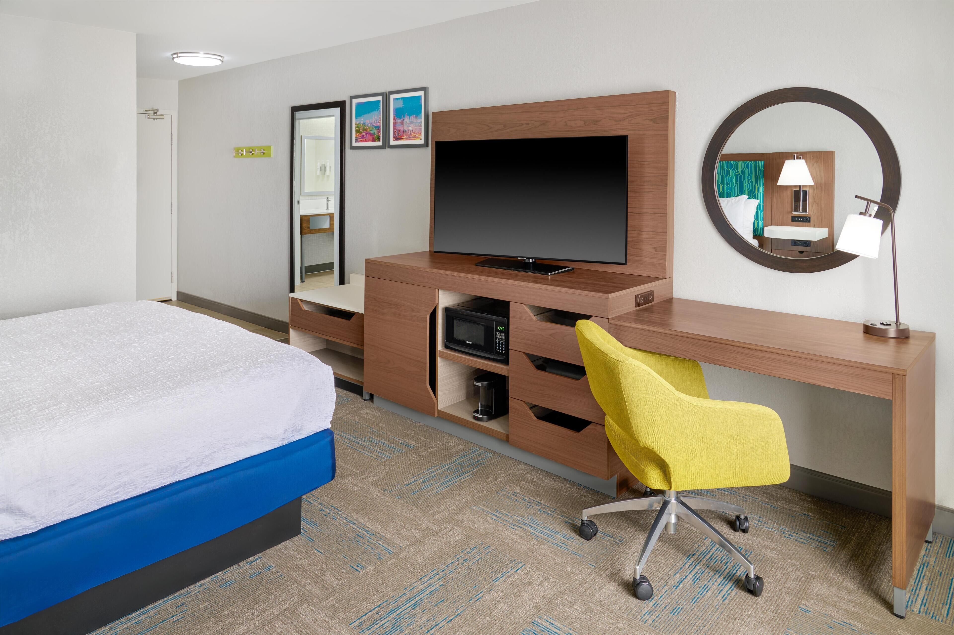 A hotel room with a bed, a desk with a TV, a chair, a mirror, and a small table with a lamp.