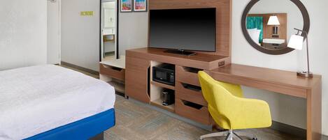 A hotel room with a bed, a desk with a TV, a chair, a mirror, and a small table with a lamp.
