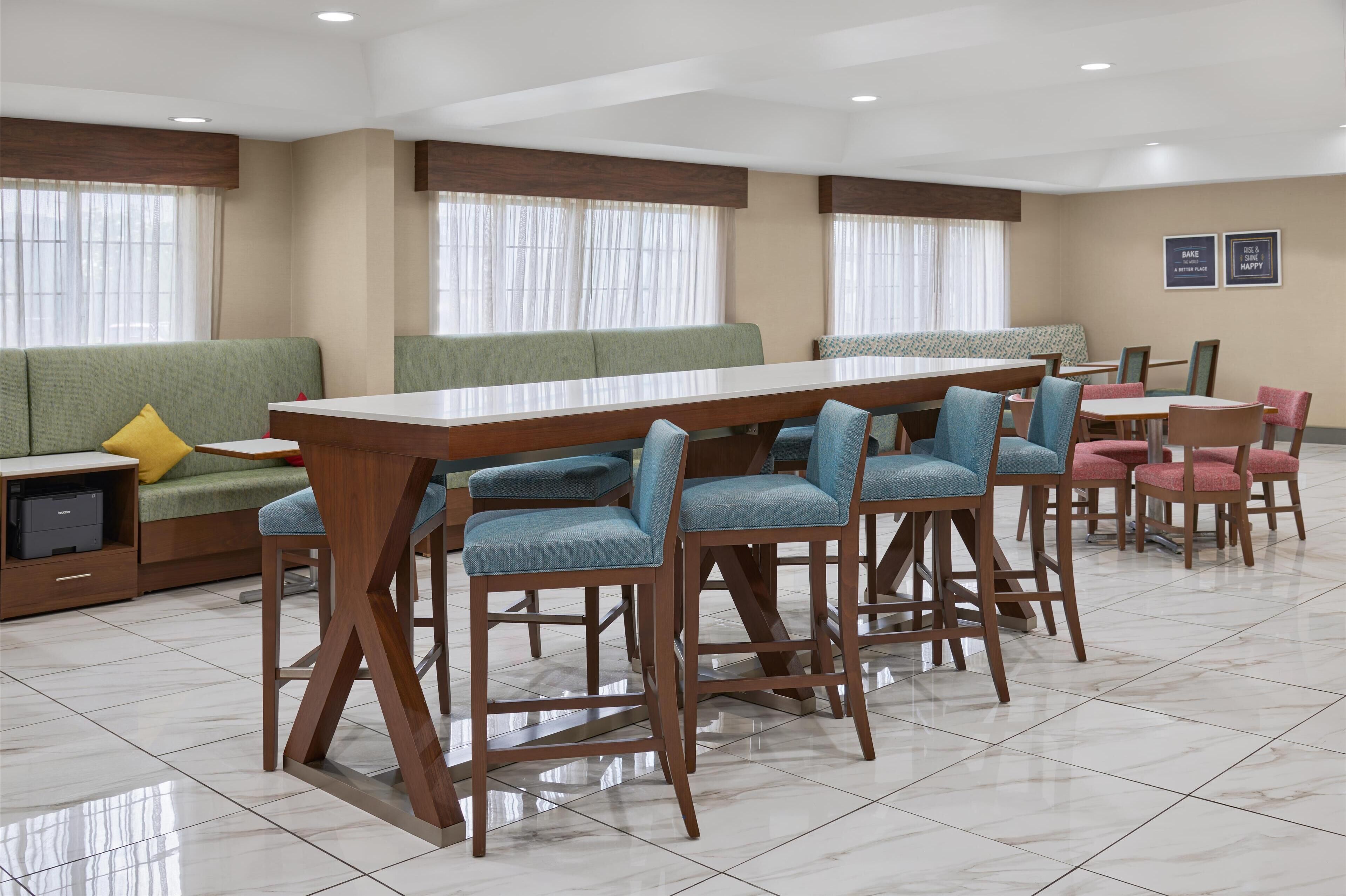 Un comedor de hotel moderno con una larga mesa de madera, sillas con cojines azules y un sofá verde con un cojín amarillo.