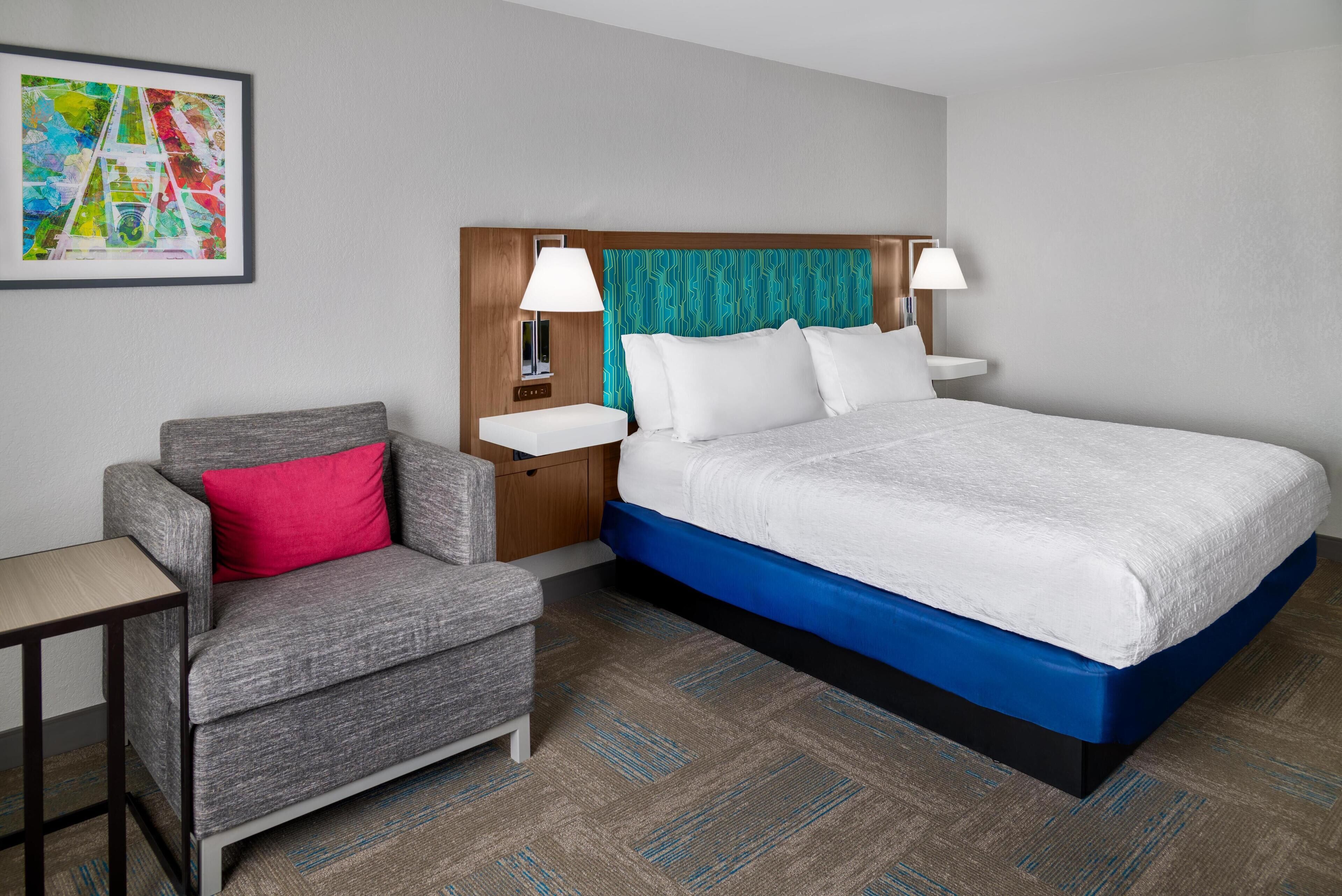 A hotel room with a bed, a grey armchair with a red pillow, a small table, and a nightstand with lamps.