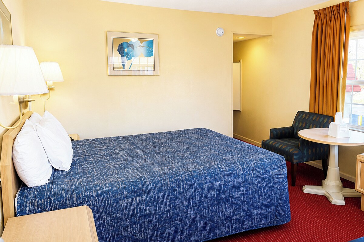 Habitación de hotel con cama, una silla, una mesita, lámpara, un cuadro y ventana con cortinas.