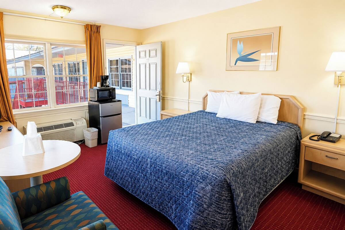 A hotel room with a bed, a desk, a television, a kitchenette, and a view of the outside.