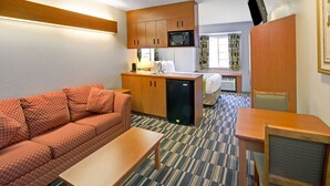A compact hotel room with a kitchenette, a sofa, a dining table, and a TV mounted on the wall.