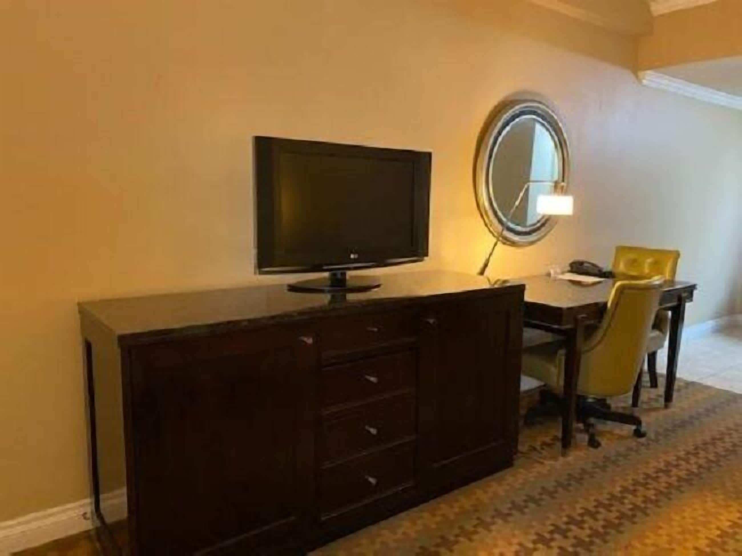 A hotel room with a dark wooden desk, a flat-screen TV, a round mirror, and a yellow chair.
