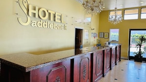 A hotel reception desk with a chandelier, a sign reading "HOTEL addleback," and a view of the outside through large windows. - Hotel Saddleback Los Angeles - Norwalk (Norwalk)