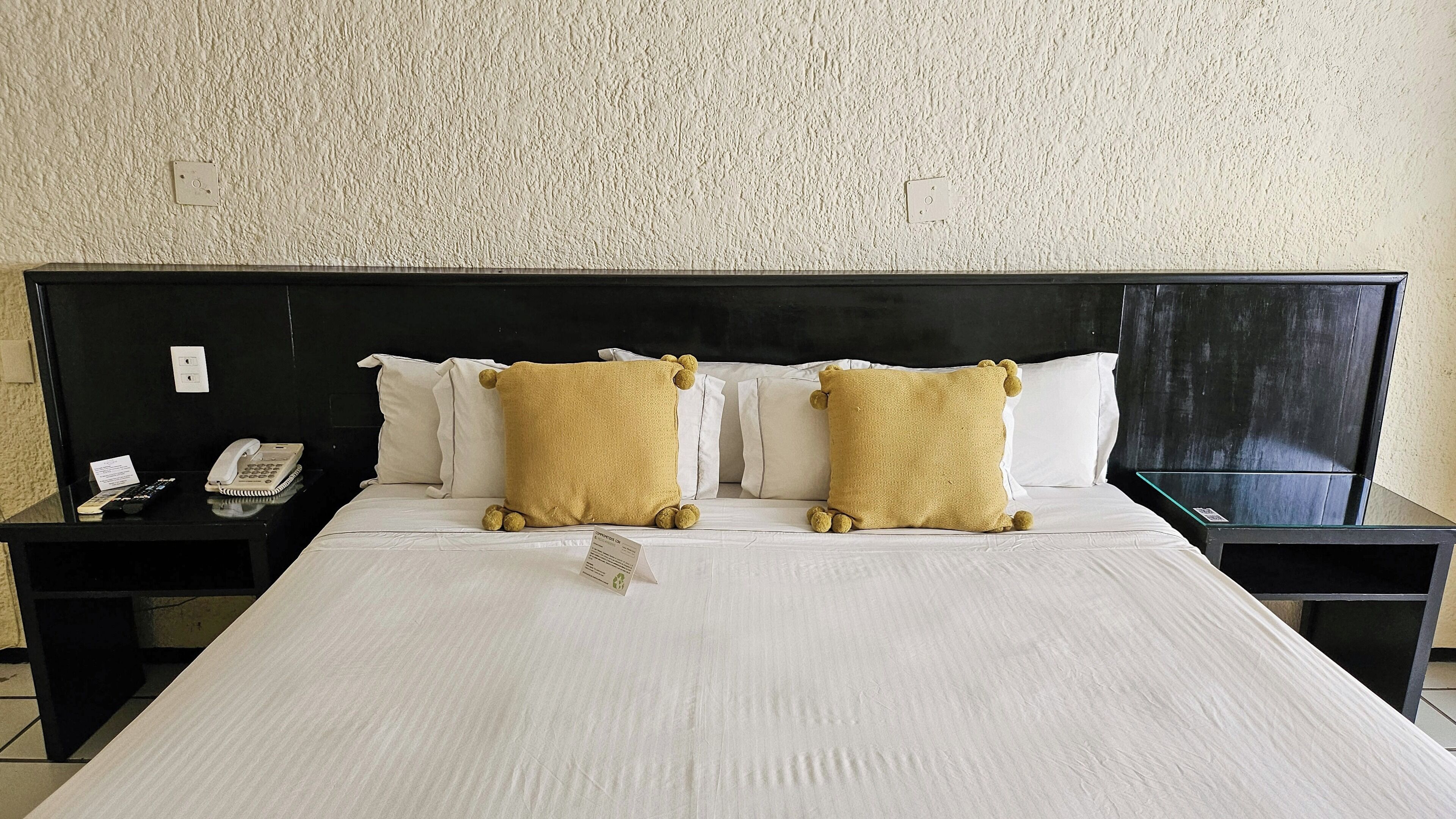 A hotel room with a large bed, two bedside tables, a telephone, and a sign on the table.