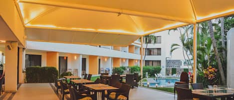 An outdoor dining area with tables and chairs, a pool in the background, and a covered walkway.