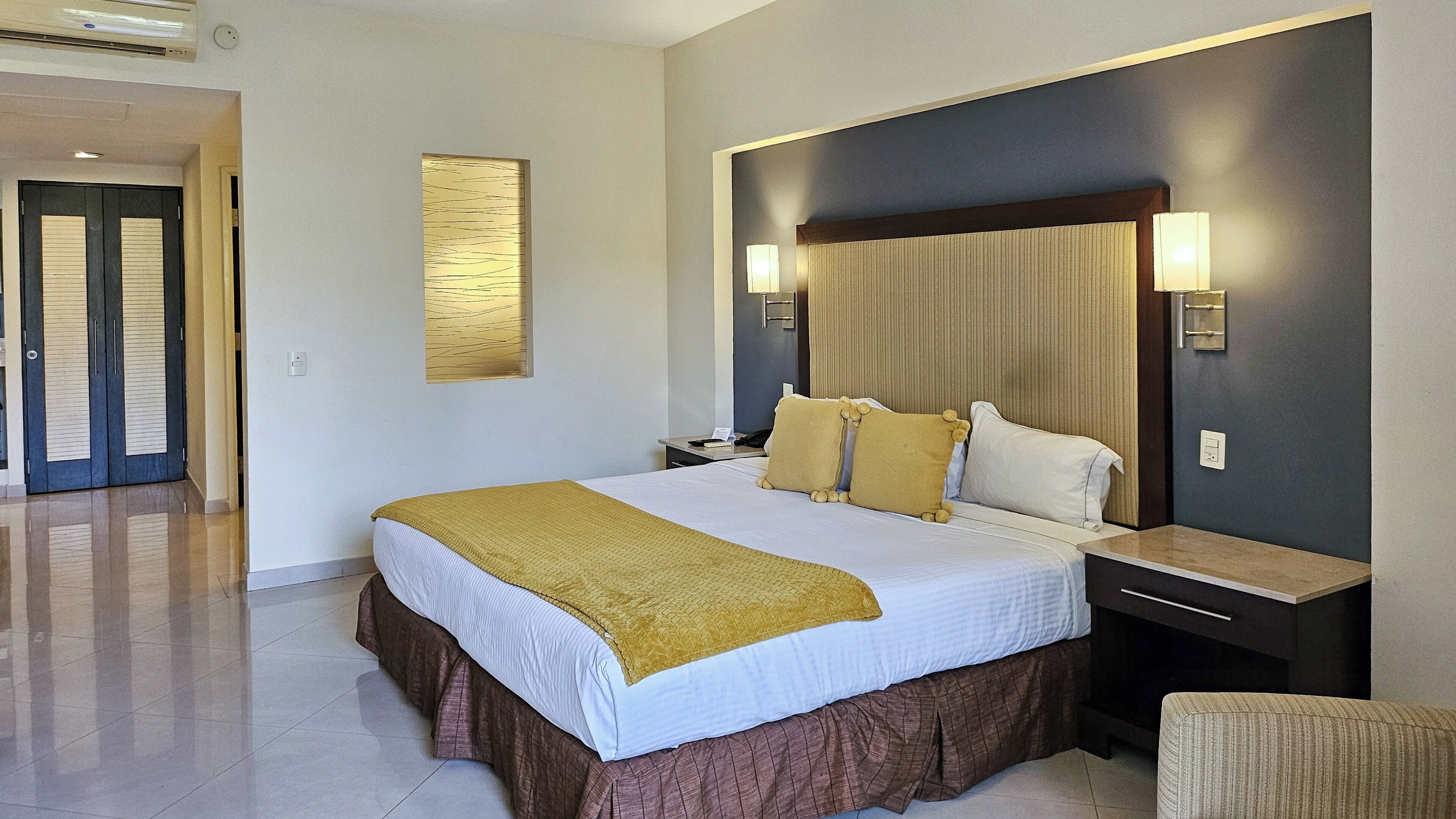 A hotel room with a large bed, a nightstand, a wall-mounted lamp, and a door leading to another room.