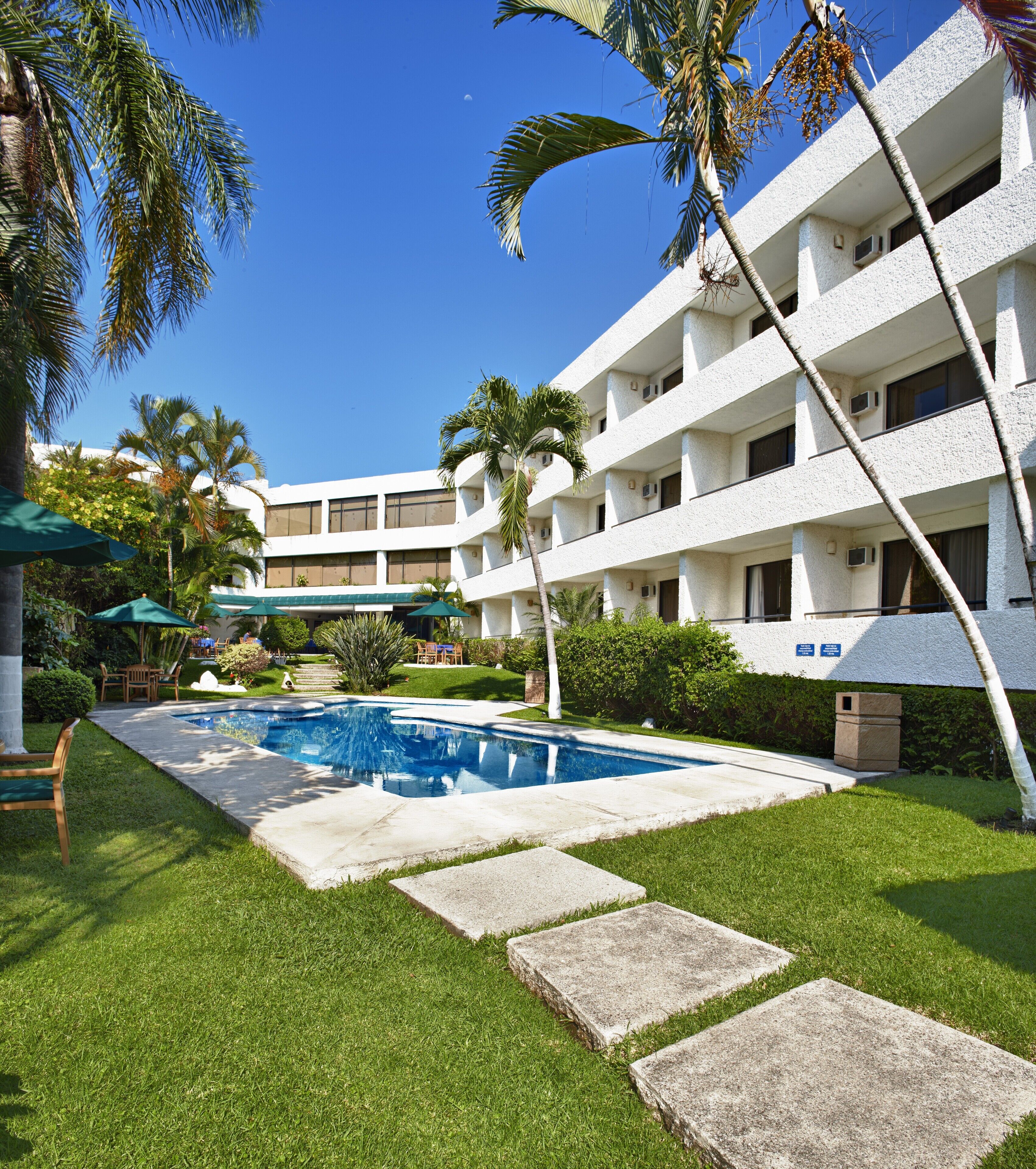A hotel with a swimming pool, palm trees, and a well-maintained lawn.