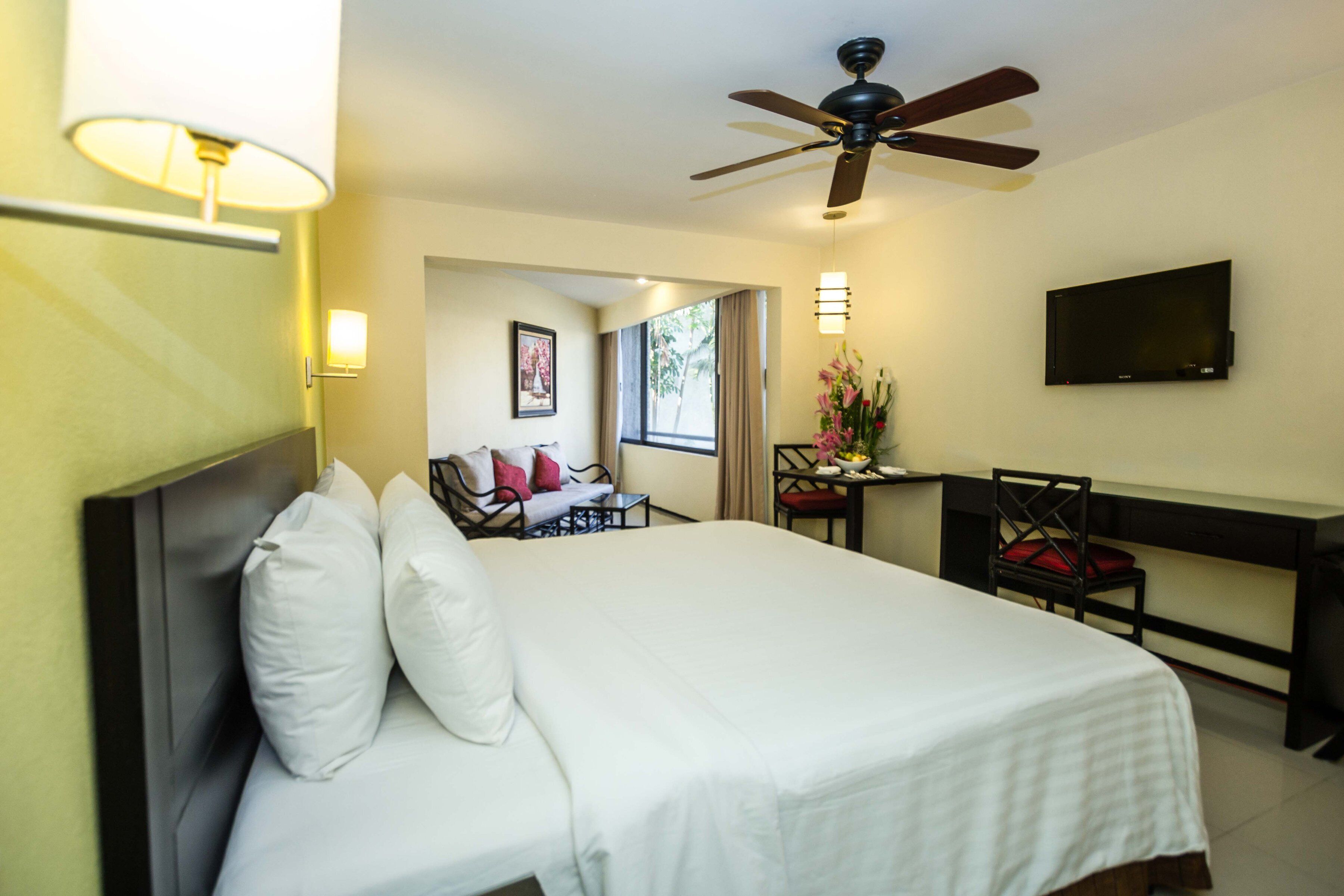 Habitación de hotel con una cama grande, un escritorio, una silla, un ventilador de techo y un televisor en la pared.