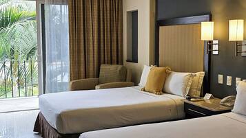 Habitación de hotel con cama, una silla, mesita de noche con teléfono y vista a palmeras a través de la ventana.