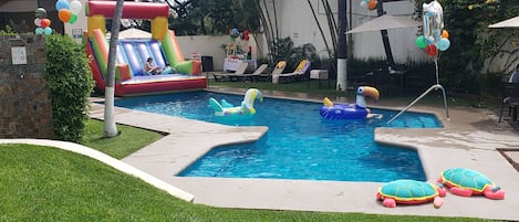 Une piscine avec des toboggans et des jeux gonflables, entourée d’une pelouse bien entretenue et d’un espace patio aménagé avec des chaises longues.