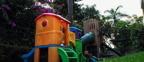 Estructura de juego infantil con tobogán y elementos para escalar, rodeada de abundante vegetación y plantas con flores.