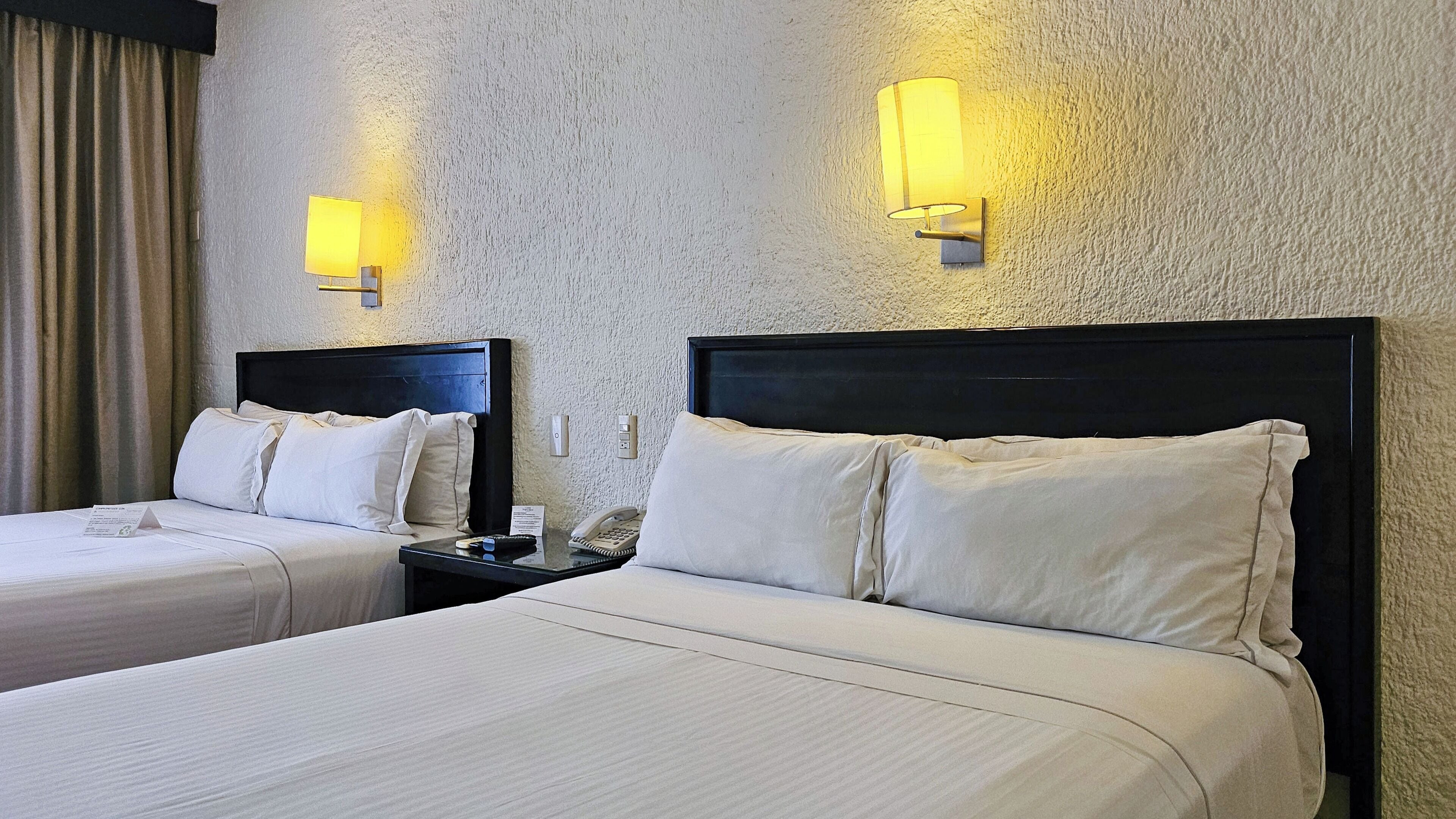 Une chambre d’hôtel avec deux lits, chacun doté de draps blancs et d’une tête de lit sombre. Des lampes murales sont fixées de chaque côté des lits.