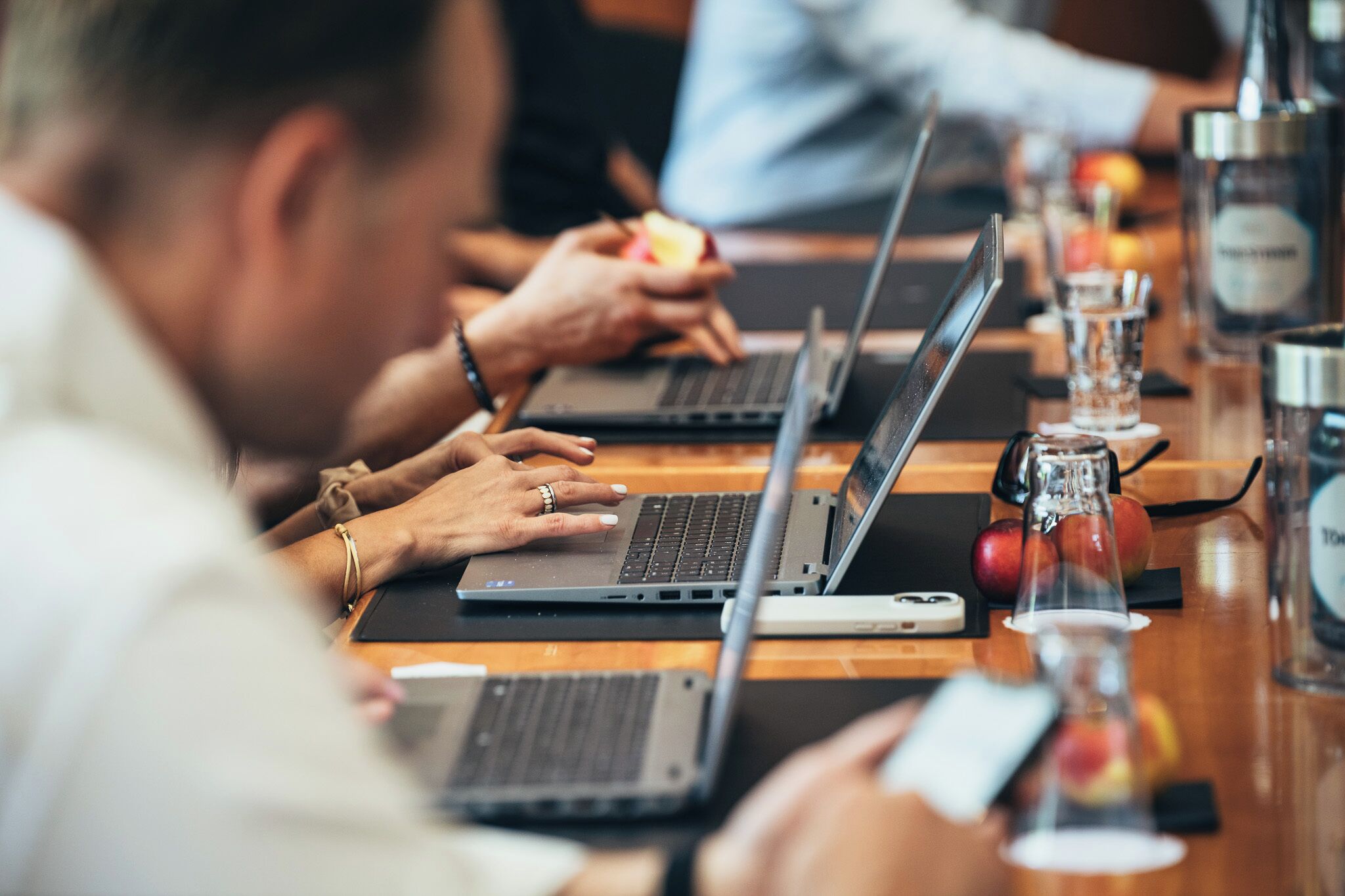 Een groep mensen werkt op laptops aan een houten tafel met drankjes en fruit.